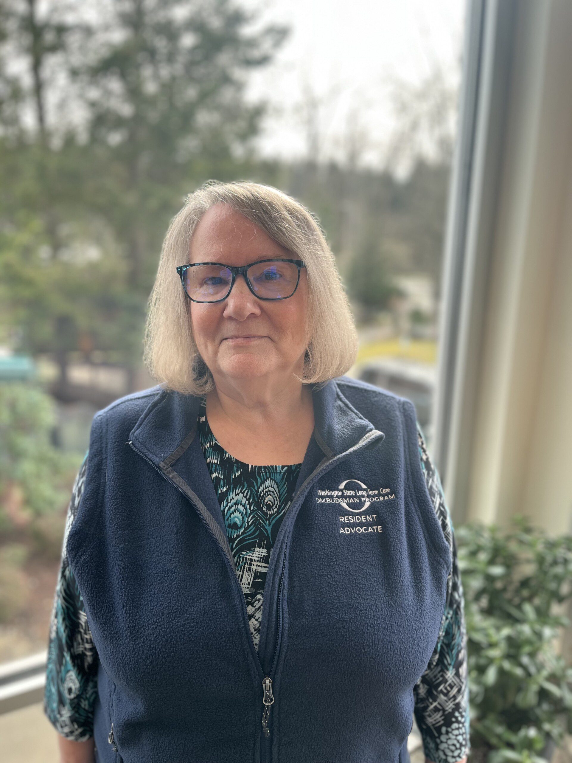 Woman with glasses, wearing a vest, smiles indoors.