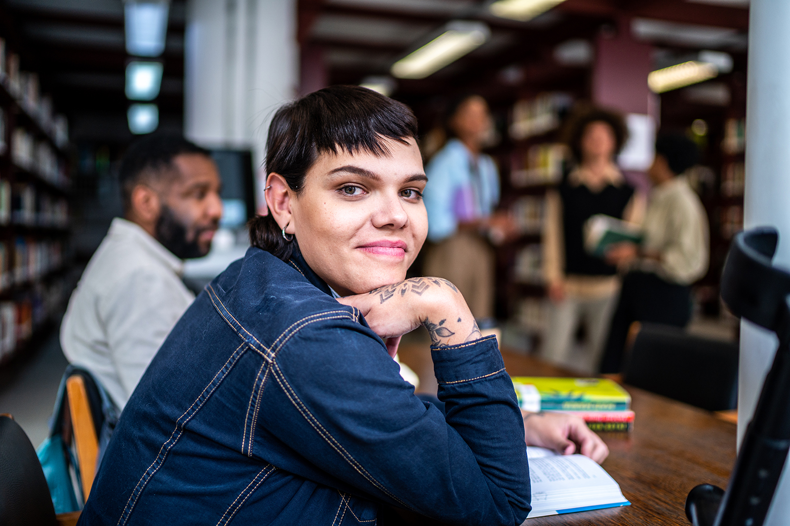 Person with tattoos in library, others interacting in background.