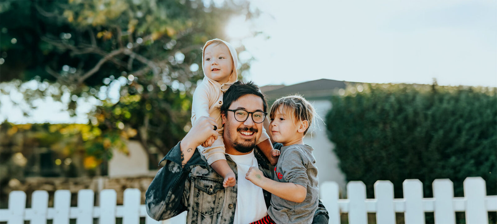 Father smiling with two children outside by a white fence.