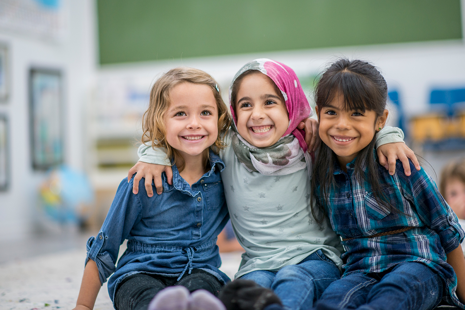 Three smiling girls embracing in a classroom setting.