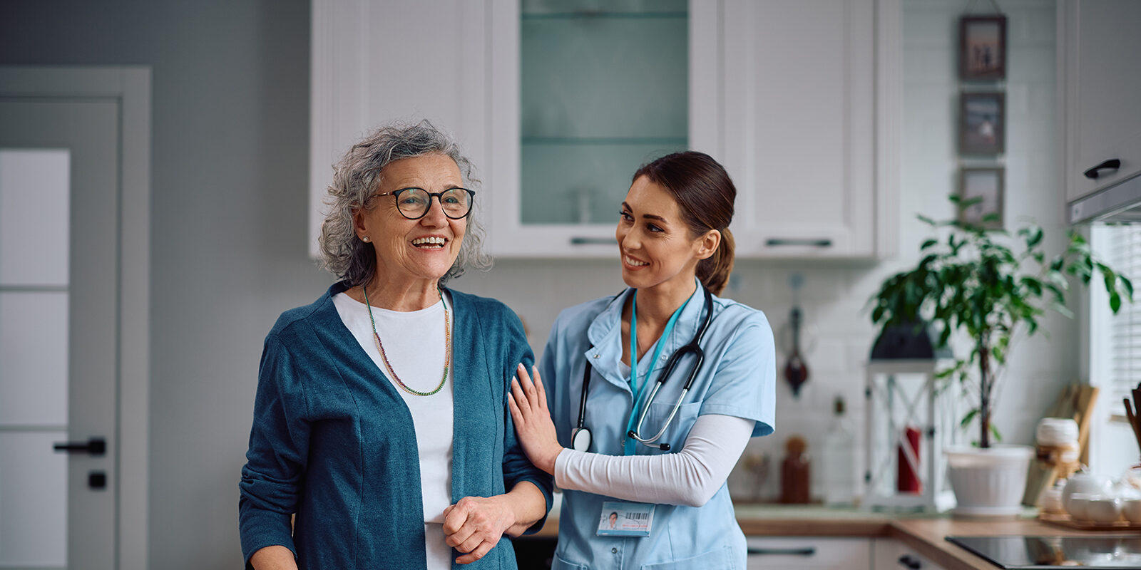 Nurse and elderly woman smiling together in a kitchen.